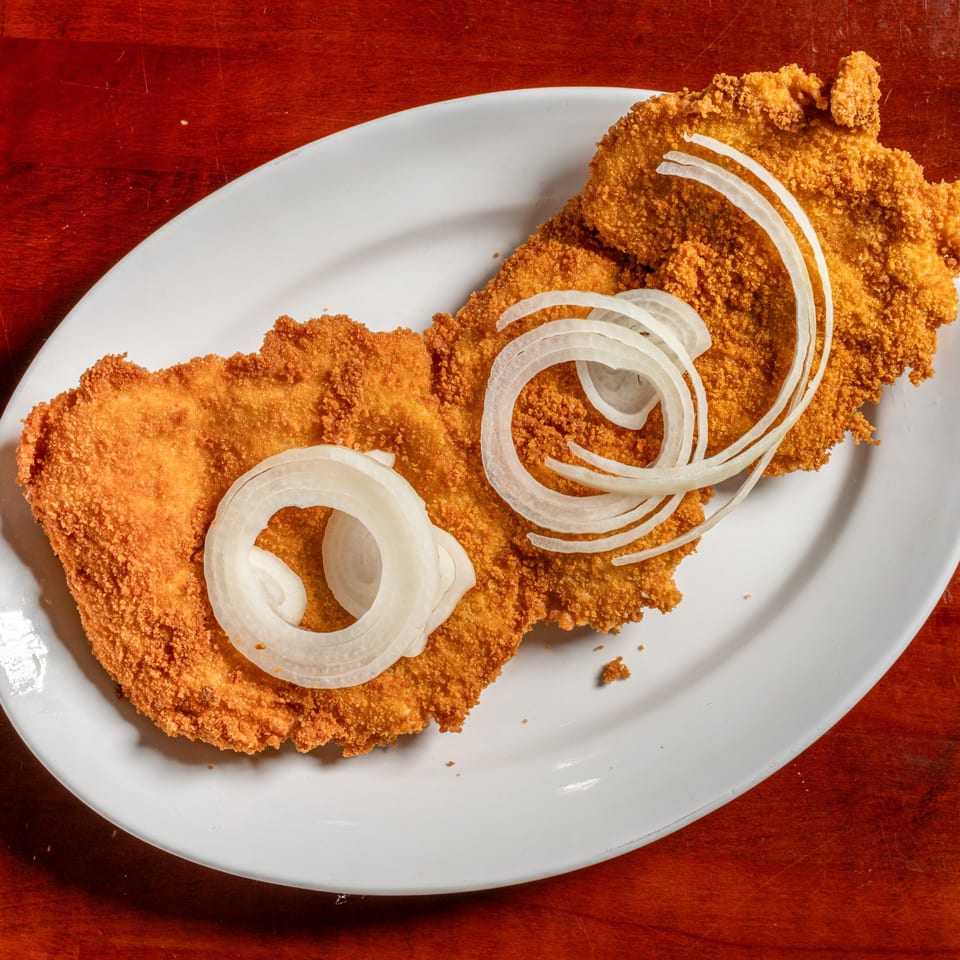 Crispy breaded Cuban steak, milanesa, topped with fresh white onion rings on a white plate.