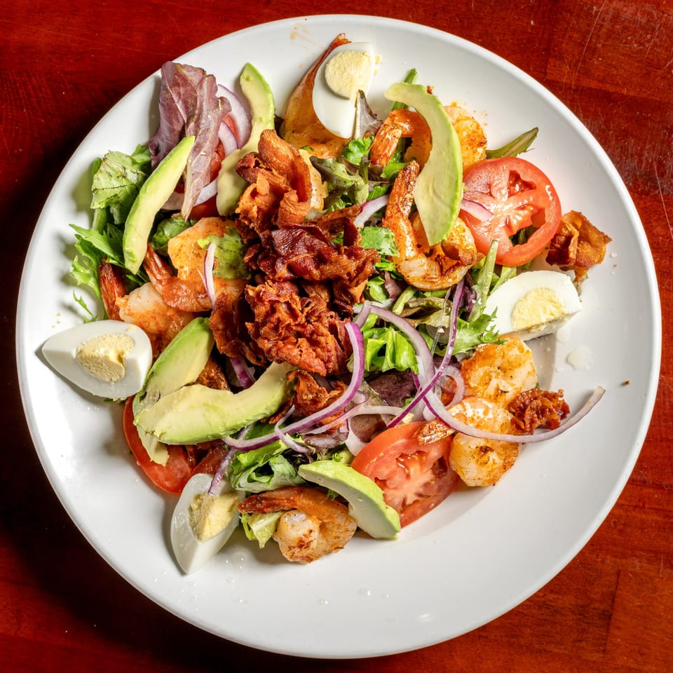 Shrimp salad with avocado, bacon, tomatoes, red onion, and hard-boiled eggs on a white plate.